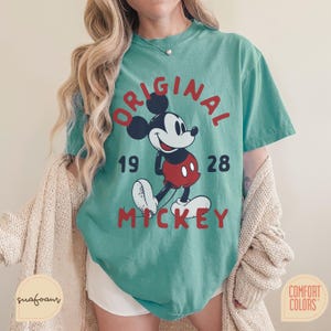 May include: Teal-colored t-shirt featuring a graphic of Mickey Mouse with the text "ORIGINAL MICKEY 1928" in red. The shirt has a vintage, slightly faded look. The shirt is made by Comfort Colors.