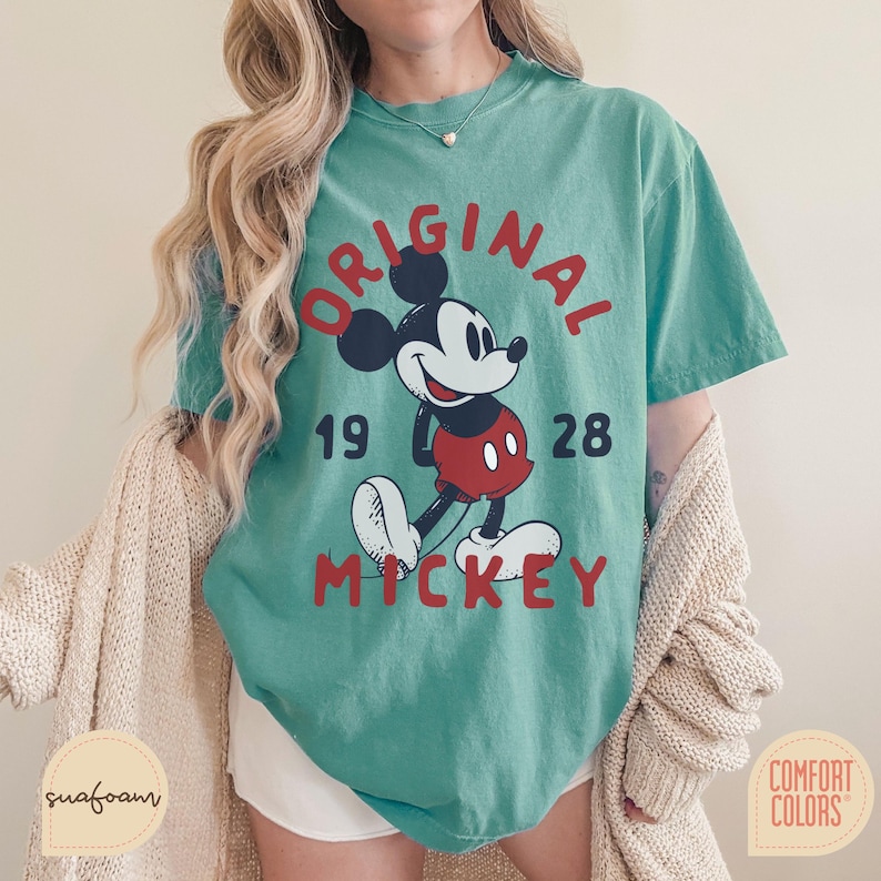 May include: Teal-colored t-shirt featuring a graphic of Mickey Mouse with the text "ORIGINAL MICKEY 1928" in red. The shirt has a vintage, slightly faded look. The shirt is made by Comfort Colors.