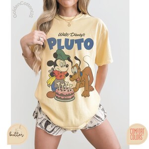 Puede incluir: Camiseta amarilla clara con un gr&aacute;fico de Mickey Mouse y Pluto, y el texto "Walt Disney's PLUTO". Mickey Mouse lleva un sombrero verde, y Pluto es marr&oacute;n. Una tarta rosa con velas en forma de hueso est&aacute; en el centro.