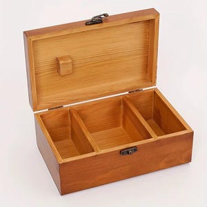 Puede incluir: Una caja de almacenamiento rectangular de madera con un acabado natural en color marrón claro y veta de madera visible. La caja cuenta con una tapa abatible con cierre metálico y el interior está dividido en tres compartimentos por dos separadores.