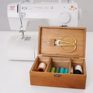 May include: A wooden sewing box with compartments, holding spools of thread in various colors and gold scissors. A white sewing machine is in the background.