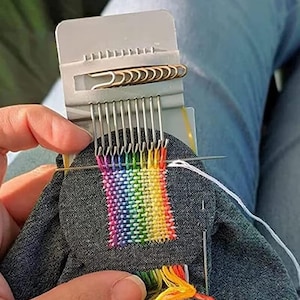 Könnte beinhalten: Ein grauer Webstuhl mit einem im Entstehen befindlichen, regenbogenfarbenen gewebten Patch. Der Patch wird mit verschiedenen farbigen Fäden hergestellt, wodurch ein lebendiges Spektrum entsteht. Eine Nadel wird in den Stoff eingeführt, und eine Büroklammer hält die Fäden.