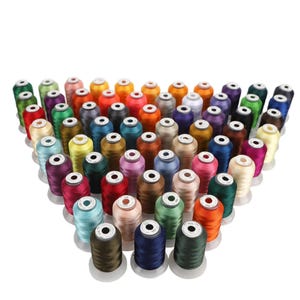 Op de afbeelding: Een grote collectie borduurgarenspoelen gerangschikt in een driehoekige formatie. De spoelen hebben een grote verscheidenheid aan levendige kleuren, waaronder rood, blauw, groen, geel, paars, bruin en wit, elk met een wit rond label bovenop.