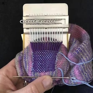 Può includere: Uno strumento per rammendare in metallo con rebbi verticali argentati e un meccanismo a scorrimento, utilizzato per riparare un tessuto a maglia multicolore viola, rosa e blu. Un ago da cucito argentato con filo azzurro è posizionato sotto il telaio.