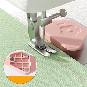 Könnte beinhalten: Eine hellrosa magnetische Nahtführung mit blumenförmigem Design, platziert neben einem silbernen Nähmaschinenfuß auf hellgrünem Stoff. Die Unterseite der Führung zeigt drei kreisförmige Magnete und eine gitterartige Struktur.