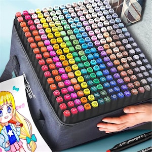 May include: A large set of colorful art markers in a black carrying case. The markers are arranged in a grid, with a wide range of colors visible. A drawing of an anime character is in the foreground. Two markers are visible in the bottom right.