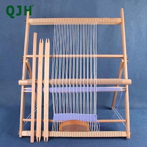 Puede incluir: Un telar de madera con hilos verticales blancos y una sección lavanda parcialmente tejida. El telar incluye herramientas de madera como un peine y lanzaderas. El marco es de madera clara, y el diseño general es simple y funcional.