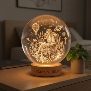 May include: A glowing glass sphere night light with a wooden base. The sphere features an engraved scene of a woman reading, surrounded by flowers, a hot air balloon, and celestial elements. The base reads "EMMA'S WORLD".