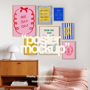 May include: A collection of framed art prints with colorful designs and text, including phrases like "Que Sera Sera" and "Meow-cha Time!" The prints are displayed on a white wall above a wooden cabinet and a pink armchair.