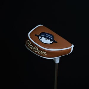 Puede incluir: Una funda para la cabeza del palo de golf marrón con un borde blanco y la palabra "Malbon" bordada en dorado. La funda presenta una bola de golf de dibujos animados con un sombrero. El fondo es negro.