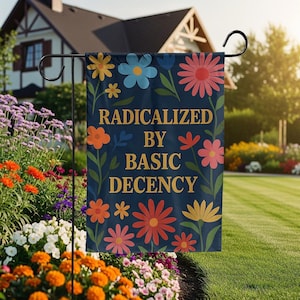 以下が含まれることがあります： 紺色のガーデンフラッグ。カラフルな花の模様と、金色の文字で「RADICALIZED BY BASIC DECENCY」と書かれています。庭に飾られており、様々な花が咲いています。