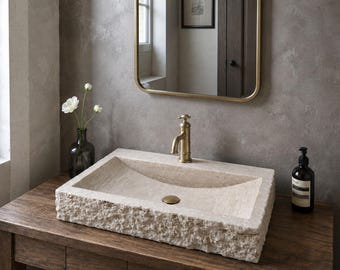 May include: A rectangular stone vessel sink with a rough, chiseled exterior and a smooth, sloped interior basin. The sink features a central brass drain and is paired with a matching brass faucet, set upon a dark wooden vanity cabinet.