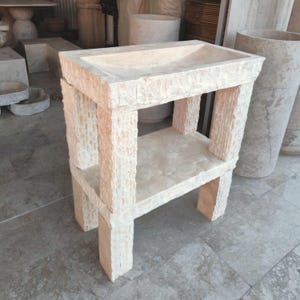Puede incluir: Un lavabo de piedra beige claro con una pila rectangular y un estante inferior. El lavabo está sostenido por cuatro columnas cuadradas texturizadas. La piedra tiene un acabado natural rugoso, y el diseño es minimalista y robusto.
