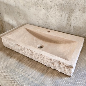 Peut inclure: Un évier rectangulaire en pierre beige avec un bassin sculpté et deux trous pour robinets. L'évier a un bord rugueux et texturé et est en pierre naturelle. L'évier est conçu pour une salle de bain.