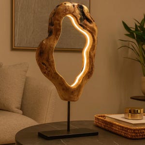 May include: A unique wooden table lamp with a natural, organic shape. The lamp features a light-colored, open-center design with a warm, glowing light. It stands on a black metal stand and base, adding a modern touch to the natural wood.
