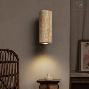 May include: A cylindrical wall sconce made of beige stone, emitting a warm light downwards. The sconce is mounted on a gray wall, with a small table and chair visible in the foreground. The sconce is a decorative lighting fixture.