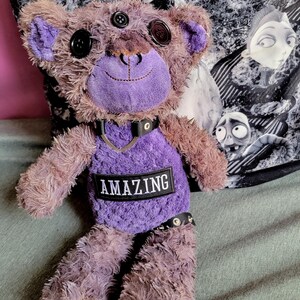 Könnte beinhalten: Ein Plüschteddybär mit lila und braunem Fell, Knopfaugen und einem aufgestickten Lächeln. Er trägt einen schwarzen Kragen mit Nieten und einen Aufnäher mit der Aufschrift "AMAZING". Der Bär hat lila Ohren und einen lila Bauch.