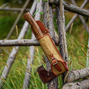 May include: A brown axe with a wooden handle is secured in a tan and brown leather sheath. The sheath has a buckle closure and a shoulder strap. The axe is hanging on a wooden fence in a natural outdoor setting.