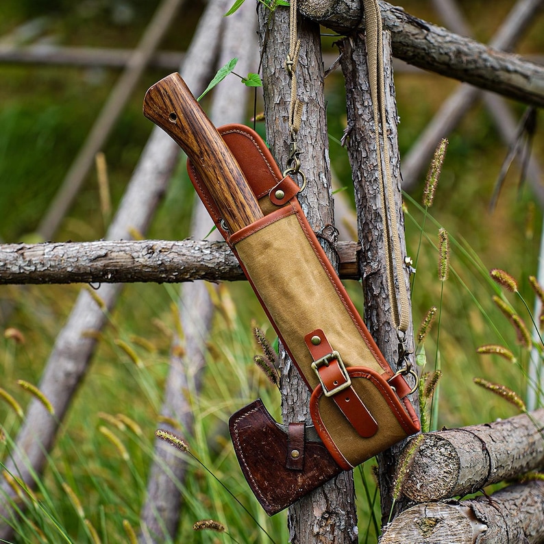 May include: A brown axe with a wooden handle is secured in a tan and brown leather sheath. The sheath has a buckle closure and a shoulder strap. The axe is hanging on a wooden fence in a natural outdoor setting.