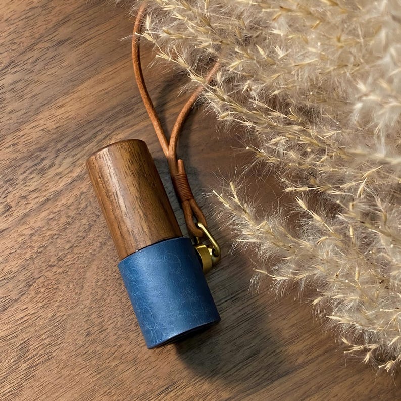 May include: A wooden and blue leather bottle stopper with a leather strap and gold-colored clasp. The top portion is made of dark wood, while the bottom is a cylinder of blue leather. The stopper is designed to seal a bottle.