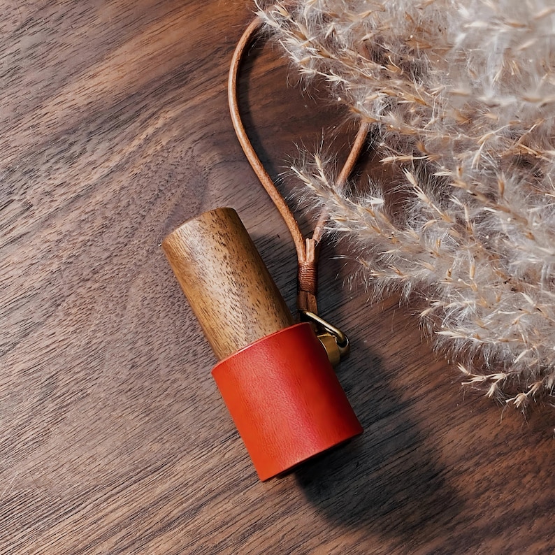 May include: A wooden bottle stopper with a vibrant orange leather band. The stopper is attached to a tan leather loop with a gold-colored clasp. The background features a wooden surface and dried ornamental grass.