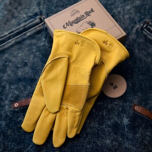 May include: A pair of yellow leather work gloves with reinforced palms and cuffs. The gloves are next to a small, round, tan-colored item and a cardboard box with the words "Adventure Rock" and a graphic of a ram.