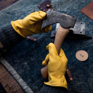 May include: Axe with a wooden handle and a metal head, held by a person wearing yellow leather work gloves. The axe head has the word "DUNHUANG" stamped on it. A small, round, tan-colored object with Japanese text is also visible.