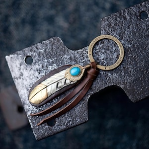 Peut inclure: Un porte-clés en cuir artisanal avec un motif de plume argentée et dorée, une pierre turquoise et des glands en cuir marron. Le porte-clés a un anneau en laiton avec le texte "MOUNTAIN ROCK WORKSHOP HAND MADE LEATHER GOODS".