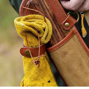 May include: Close-up of a leather quiver with a rolled yellow glove secured by a brown leather strap. The quiver has a wooden back and a canvas front with brown leather trim. A small yellow tool hangs from a brass ring.