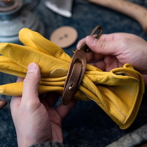 May include: A pair of bright yellow leather work gloves, held together with a brown leather strap and a metal clasp. The gloves are soft and pliable, suggesting they are well-worn. The background includes tools and a dark surface.