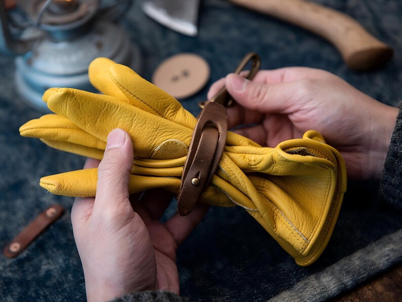 May include: A pair of bright yellow leather work gloves, held together with a brown leather strap and a metal clasp. The gloves are soft and pliable, suggesting they are well-worn. The background includes tools and a dark surface.
