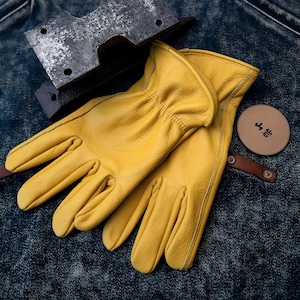 May include: A pair of bright yellow leather work gloves with white stitching. A metal tool and a round leather tag with Japanese text are also visible. The gloves are resting on a denim surface.