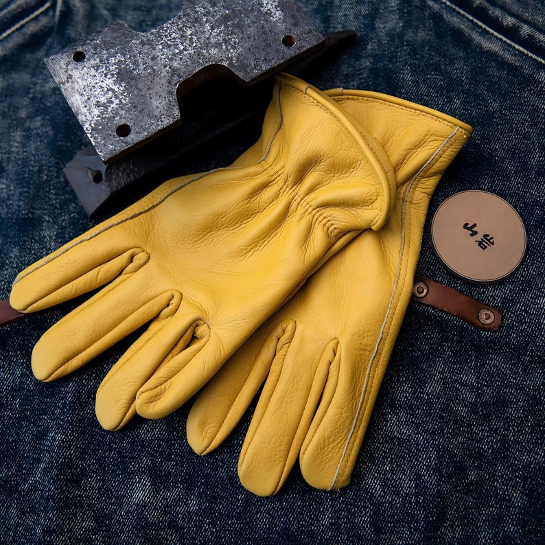 May include: A pair of bright yellow leather work gloves with white stitching. A metal tool and a round leather tag with Japanese text are also visible. The gloves are resting on a denim surface.