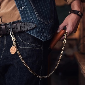 Könnte beinhalten: Eine braune Ledergeldbörse mit einer goldenen Kette, die an einer Gürtelschlaufe befestigt ist. Die Kette ist mit einem kleinen, runden, hellbraunen Lederanhänger mit einem Design verbunden. Die Person trägt eine Jeansjacke und eine dunkelblaue Jeans.