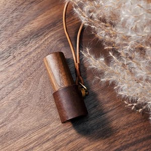 May include: A wooden and leather accessory with a brown leather band and a wooden top. The accessory has a leather strap with a gold-colored clasp. The background is a wooden surface with dried plants.