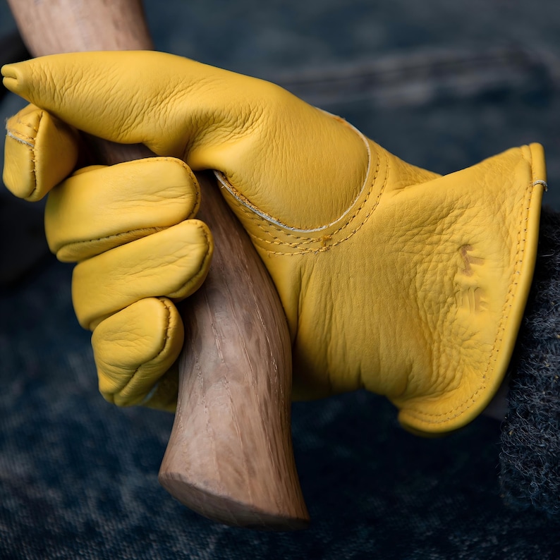 May include: Close-up of a yellow leather work glove gripping a wooden axe handle. The glove is a vibrant yellow with visible stitching and a small logo. The axe handle is made of light brown wood.