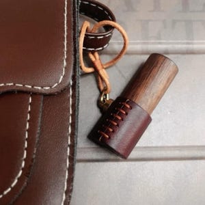 May include: A brown leather crossbody bag with white stitching is shown. A wooden object with a leather sleeve and decorative stitching hangs from the bag. The wooden object is attached to the bag with a leather strap.
