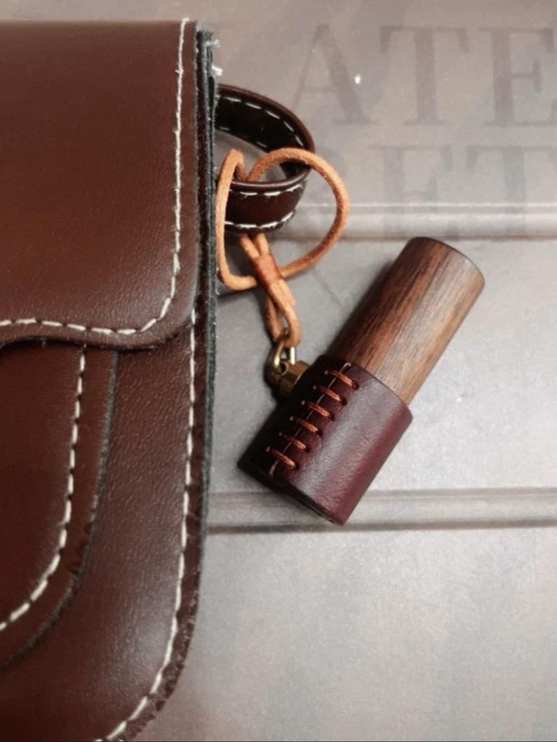 May include: A brown leather crossbody bag with white stitching is shown. A wooden object with a leather sleeve and decorative stitching hangs from the bag. The wooden object is attached to the bag with a leather strap.