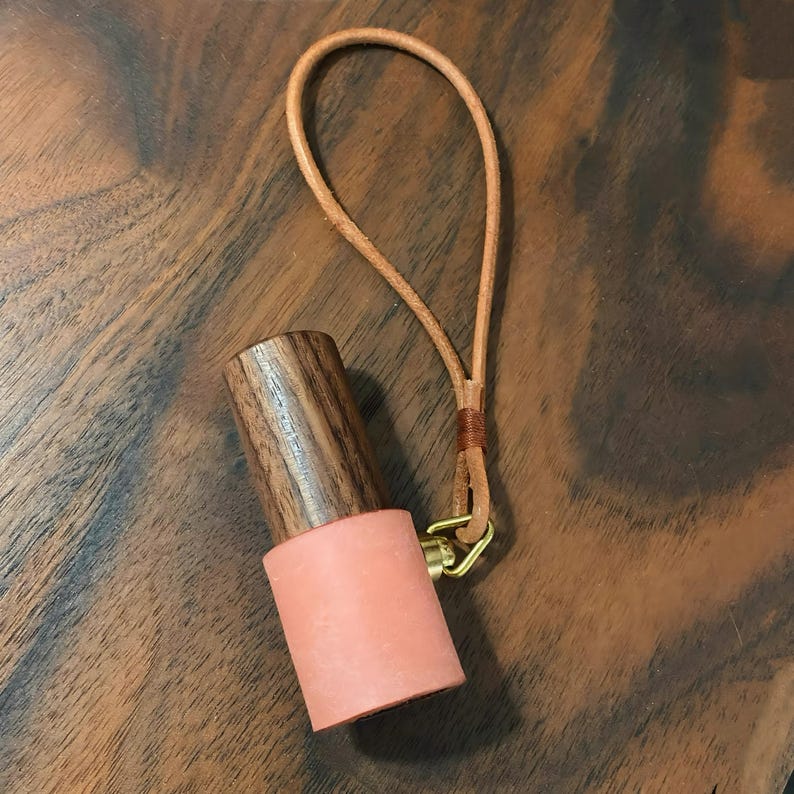 May include: A wooden and coral-colored bottle stopper with a leather loop. The top portion is made of dark wood, while the bottom is a coral-colored material. A small brass clasp connects the stopper to a tan leather loop.