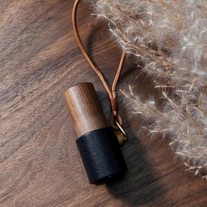 May include: A cylindrical essential oil diffuser with a dark wood top and a black leather bottom. It has a tan leather strap and a small gold-colored metal ring. The background is a wooden surface with dried plants.