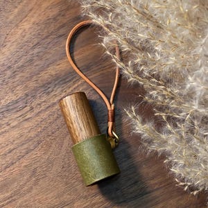May include: A wooden and olive green leather accessory with a leather loop and brass hardware. The wooden portion has a natural wood grain. The accessory is on a wooden surface with dried plants in the background.