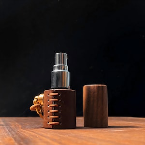 May include: A perfume atomizer with a silver metal top and a brown leather sleeve. The sleeve is stitched with orange thread. A wooden cap sits next to the atomizer. A gold-colored ring and clasp are attached to the leather sleeve.