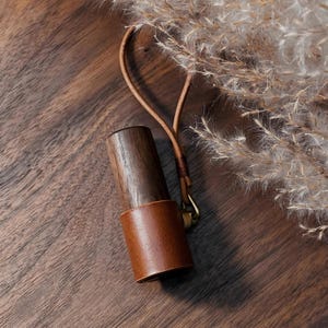 Op de afbeelding: Een houten lippenstifthouder met een bruine leren band en een leren riem. De houder is gemaakt van donker hout en heeft een messing sluiting. De achtergrond toont gedroogde planten op een houten oppervlak.