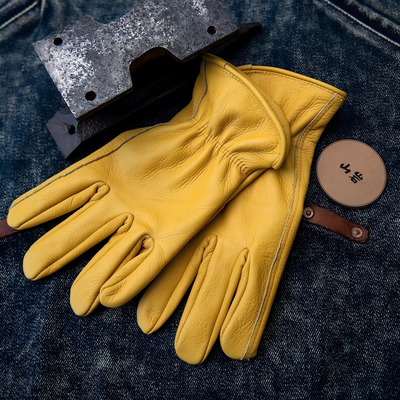 May include: A pair of bright yellow leather work gloves with white stitching. A metal tool and a round leather tag with Japanese text are also visible. The gloves are resting on a denim surface.
