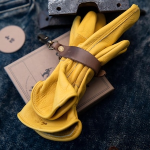May include: A pair of bright yellow leather work gloves, secured with a brown leather strap and a metal clasp. The gloves rest on a small, brown cardboard box. The background is a textured, dark blue denim.