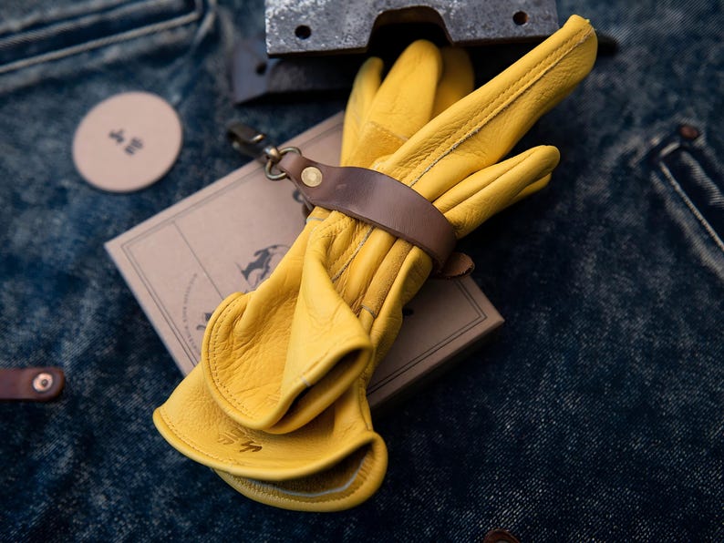 May include: A pair of bright yellow leather work gloves, secured with a brown leather strap and a metal clasp. The gloves rest on a small, brown cardboard box. The background is a textured, dark blue denim.