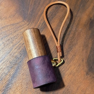 May include: A handcrafted wooden bottle stopper with a dark purple leather base and a light brown leather loop. The stopper has a brass ring and a natural wood grain finish.