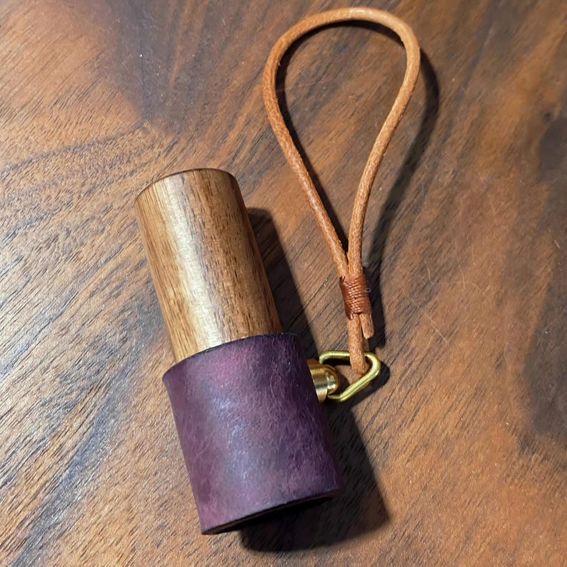 May include: A handcrafted wooden bottle stopper with a dark purple leather base and a light brown leather loop. The stopper has a brass ring and a natural wood grain finish.