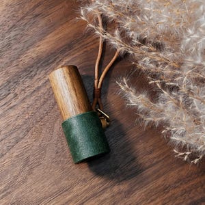 May include: A wooden and green leather bottle stopper with a gold-colored ring. The top is made of light brown wood, and the bottom is wrapped in dark green leather. The stopper is attached to a brown leather cord.