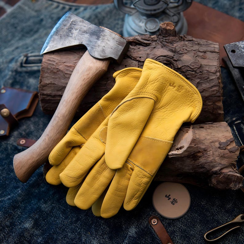 May include: Axe with a wooden handle and a metal head, resting on a piece of wood, next to a pair of yellow leather work gloves. A small, round container with Japanese text is also visible.